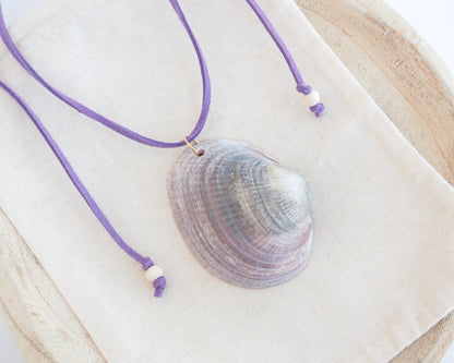 A purple cord necklace with a natural clam shell pendant, displayed on a cream fabric pouch over a light wood surface. The shell is a Violet Bittersweet Clam Shell handpicked from the beaches of Portugal.