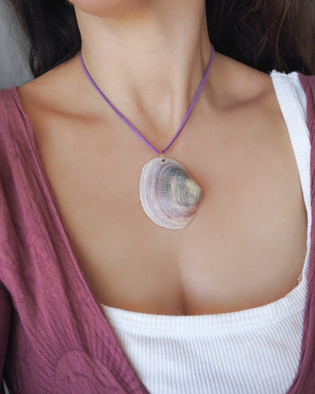 A close-up of a person wearing a purple cord necklace with a large seashell pendant, styled over a white ribbed tank top and a mauve cardigan. The shell is a Violet Bittersweet Clam Shell handpicked from the beaches of Portugal.