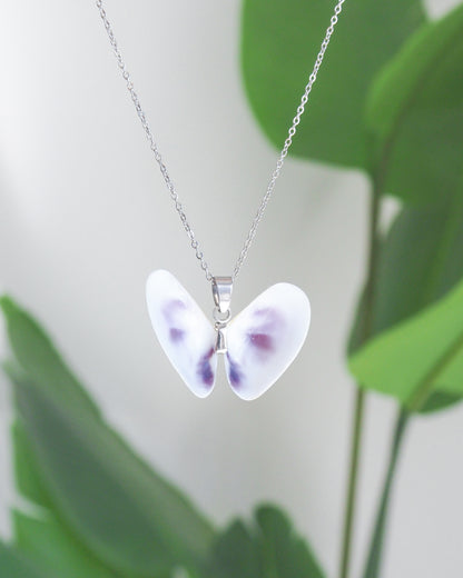 A silver chain necklace with a butterfly-shaped pendant necklace made from two white purple coquina shells, shown against a soft-focus background of green leaves.