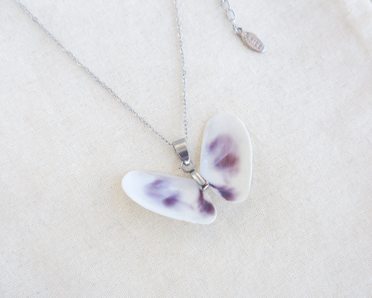 A butterfly-shaped pendant necklace made from two white purple coquina shells set on a silver chain on a textured beige linen background.