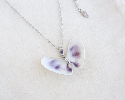 A butterfly-shaped pendant necklace made from two white purple coquina shells set on a silver chain on a textured beige linen background.