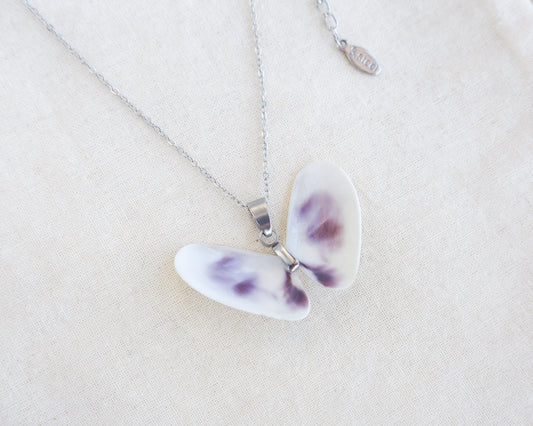 A butterfly-shaped pendant necklace made from two white purple coquina shells set on a silver chain on a textured beige linen background.