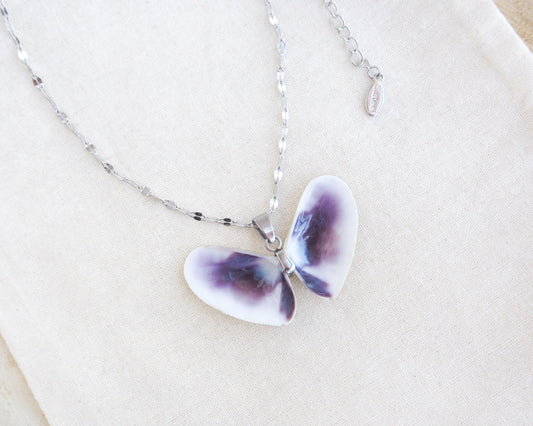 A butterfly-shaped pendant necklace made from two white purple coquina shells set on a deluxe silver chain on a textured beige linen background.