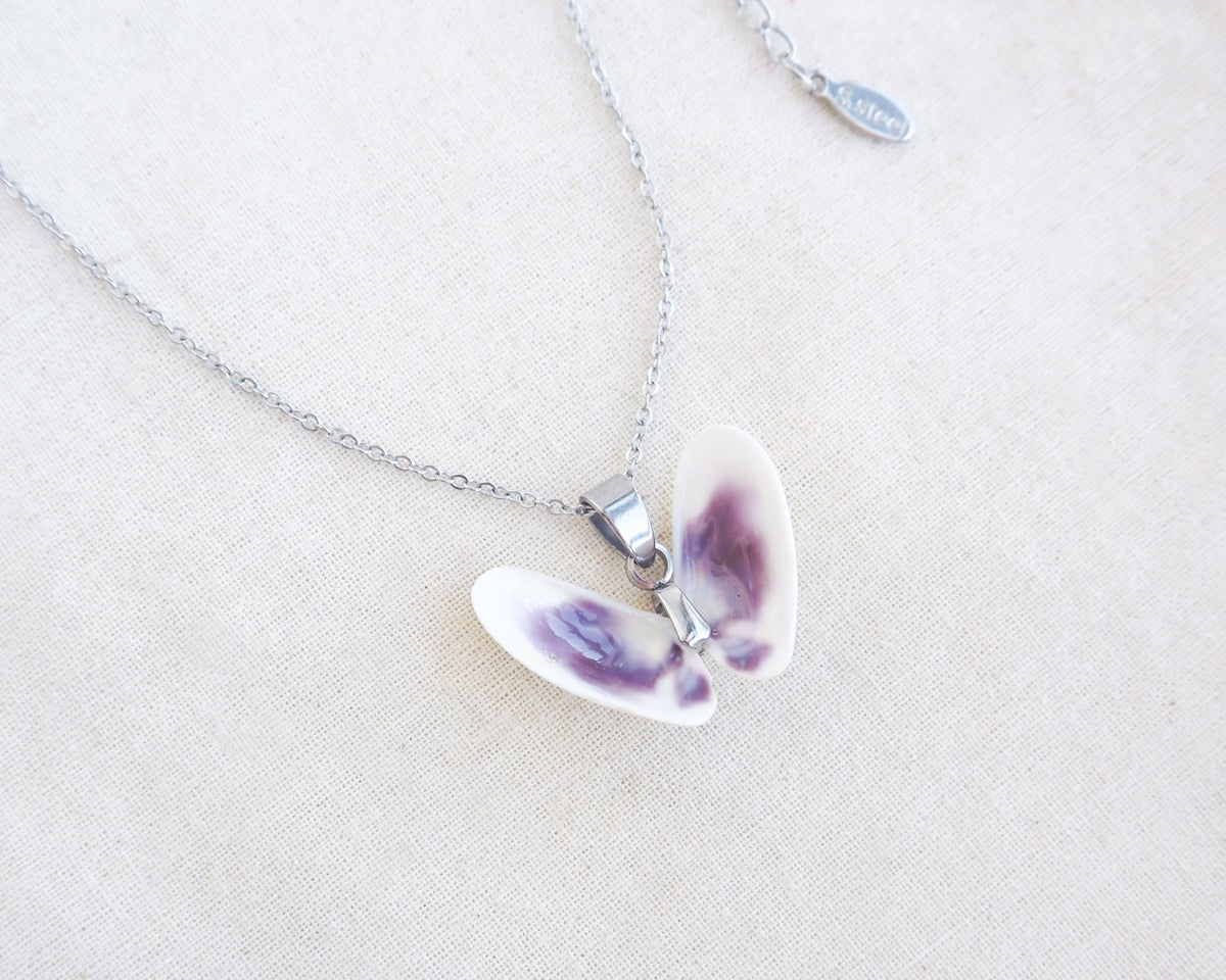 A tiny butterfly-shaped pendant necklace made from two white purple coquina shells set on a silver chain on a textured beige linen background.