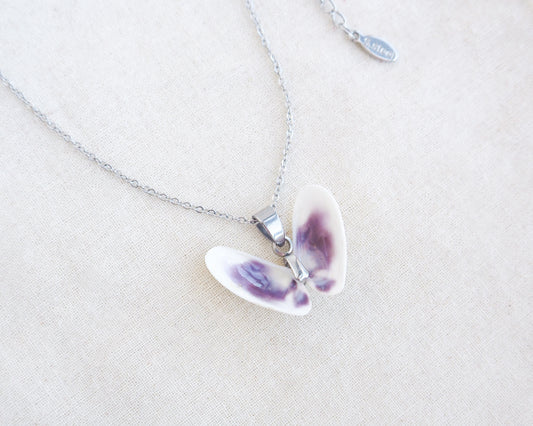 A tiny butterfly-shaped pendant necklace made from two white purple coquina shells set on a silver chain on a textured beige linen background.