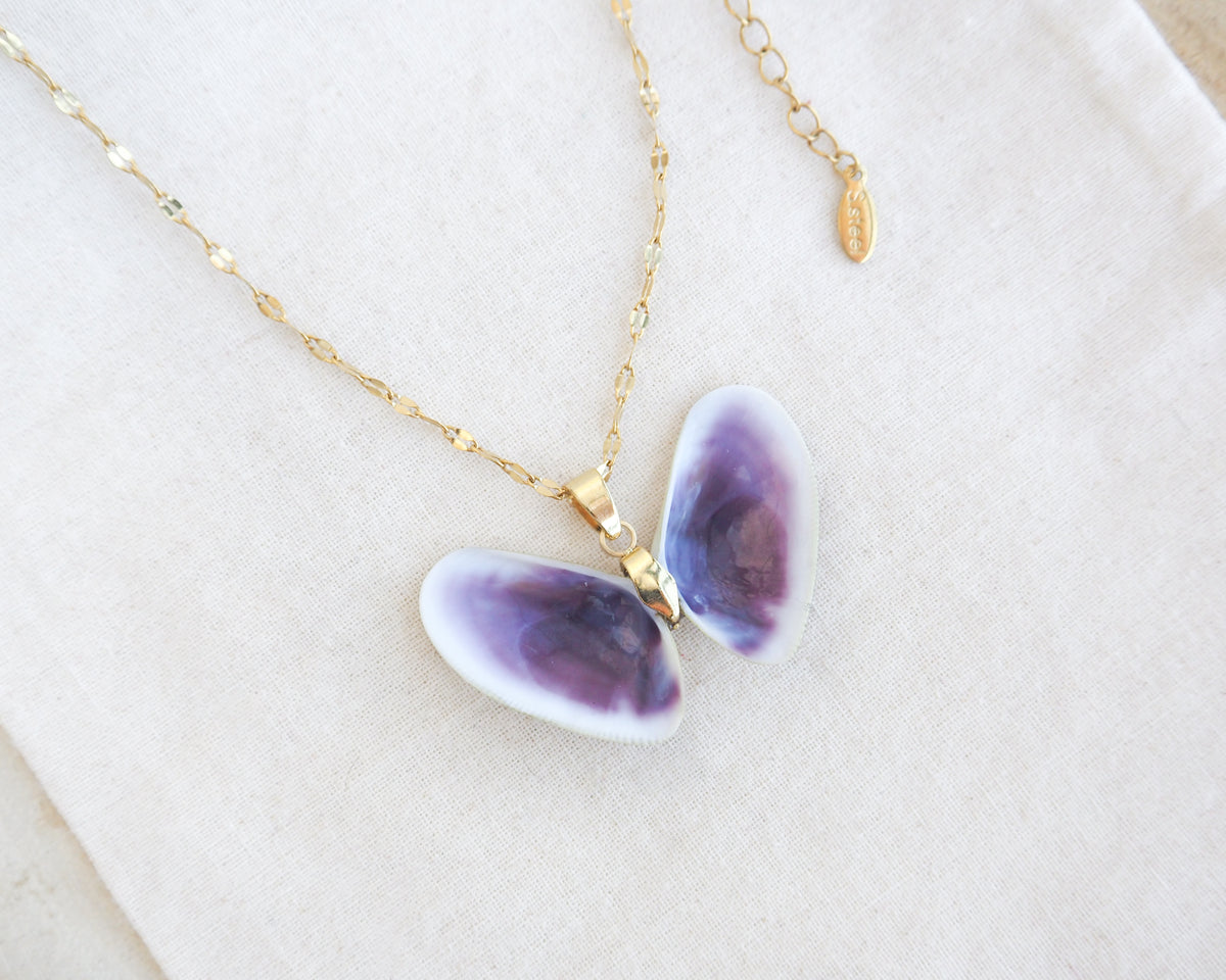 A gold chain necklace with a butterfly-shaped pendant made from two purple coquina seashells showing purple and white patterns, displayed on a textured cream fabric background.