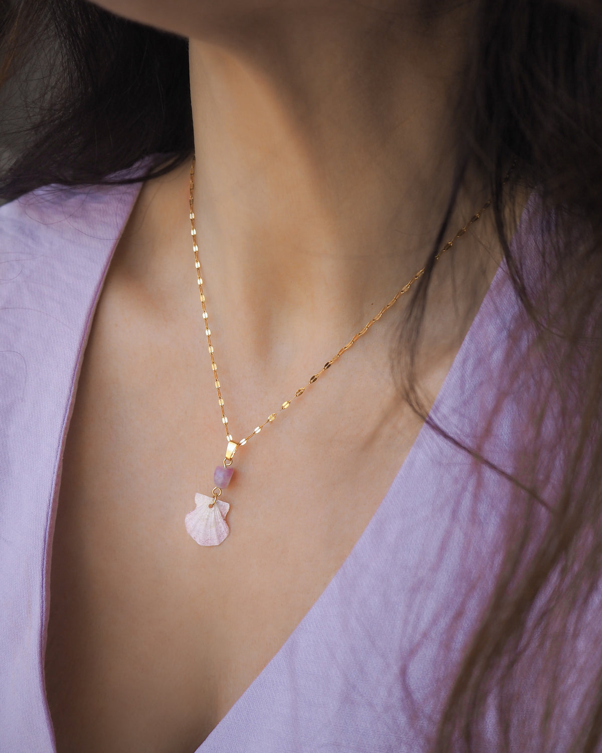 A close-up of a woman wearing a delicate gold chain necklace with a tiny soft pink scallop shell and a purple amethyst gemstone bead. The shell is a scallop shell handpicked from the beaches of Portugal and it has a soft pink color.