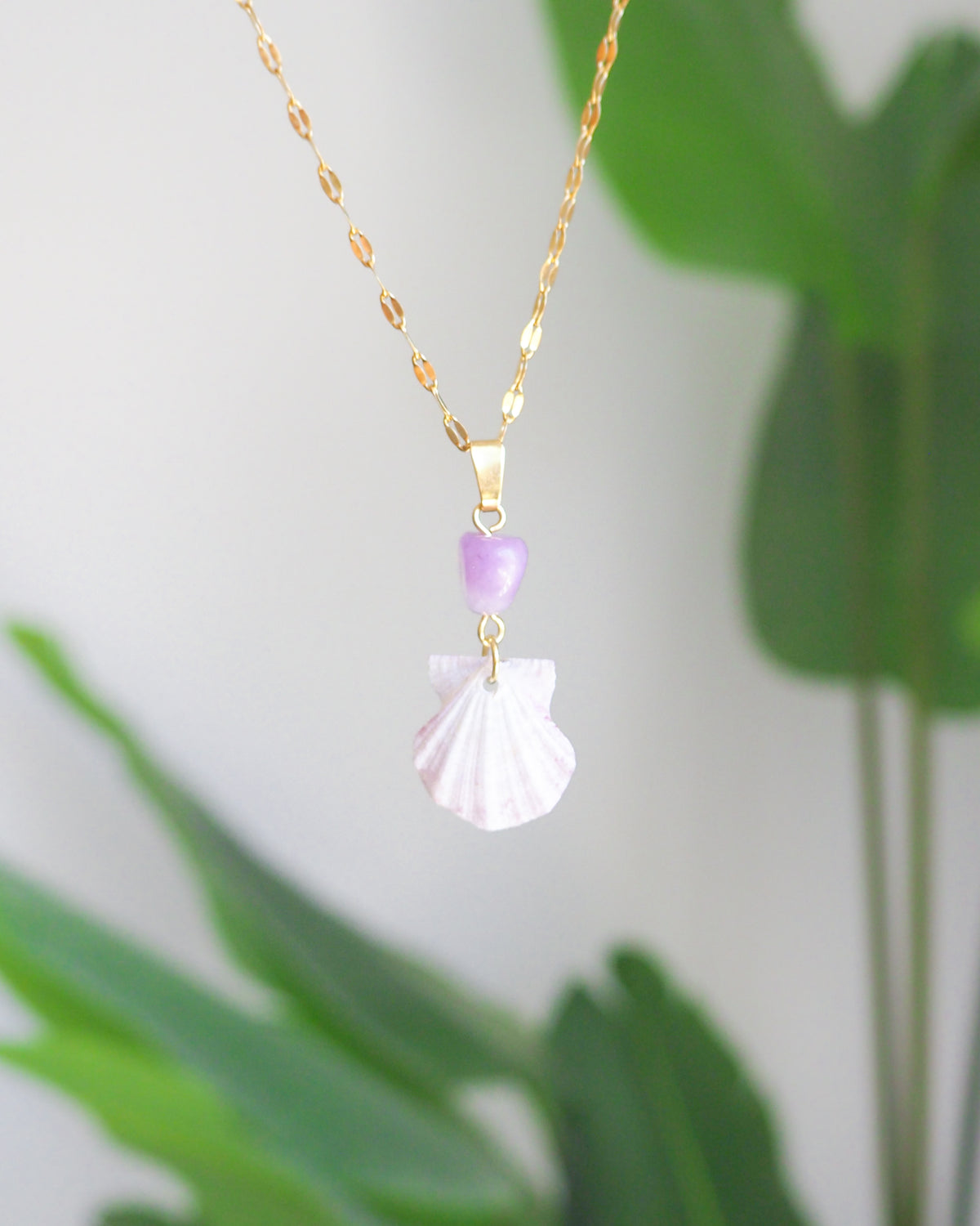 A gold chain necklace featuring a white seashell pendant and a small lavender purple bead, shown against a soft-focus background of green leaves and a light gray wall.