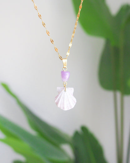 A gold chain necklace featuring a white seashell pendant and a small lavender purple bead, shown against a soft-focus background of green leaves and a light gray wall.