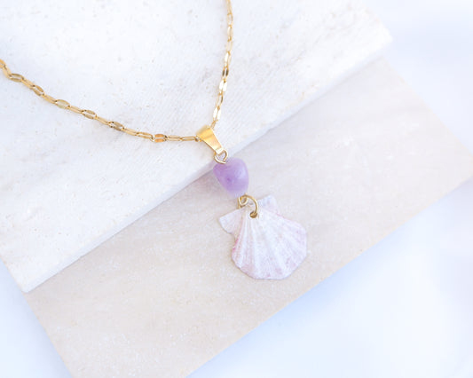 A gold chain necklace with a tiny soft pink scallop shell and a purple amethyst gemstone bead on stone displayed. The shell is a scallop shell handpicked from the beaches of Portugal and it has a soft pink color.