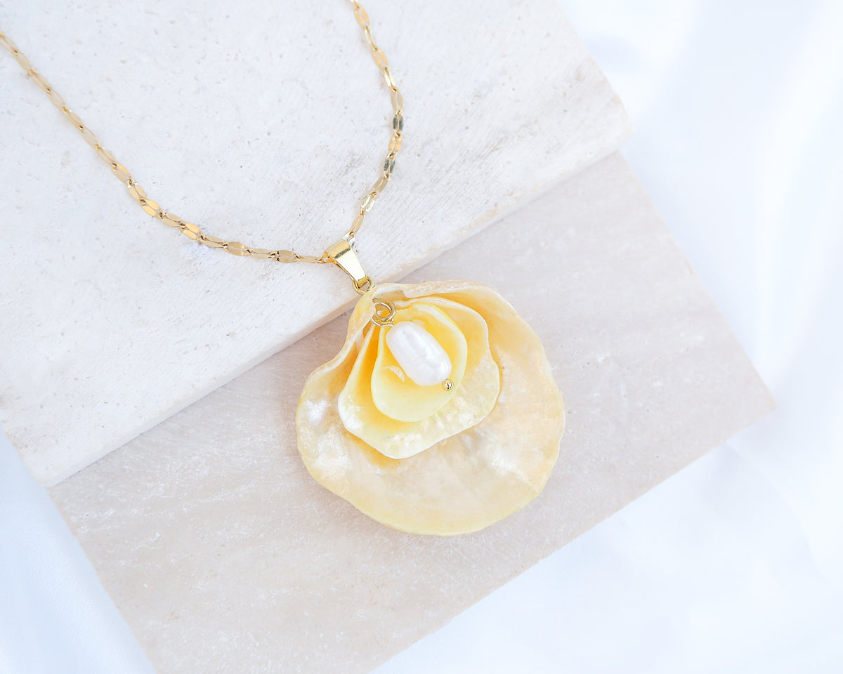 A gold chain necklace with a pendant made from layered yellow jingle shells with a layer of mother-of-pearl and a small white freshwater pearl in the center, displayed on a light textured stone block against a white fabric background. The jingle shells or saddle oyster shells are handpicked from the beaches of Portugal. Set on a gold stainless steel chain. 