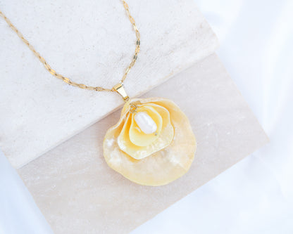 A gold chain necklace with a pendant made from layered yellow jingle shells with a layer of mother-of-pearl and a small white freshwater pearl in the center, displayed on a light textured stone block against a white fabric background. The jingle shells or saddle oyster shells are handpicked from the beaches of Portugal. Set on a gold stainless steel chain. 