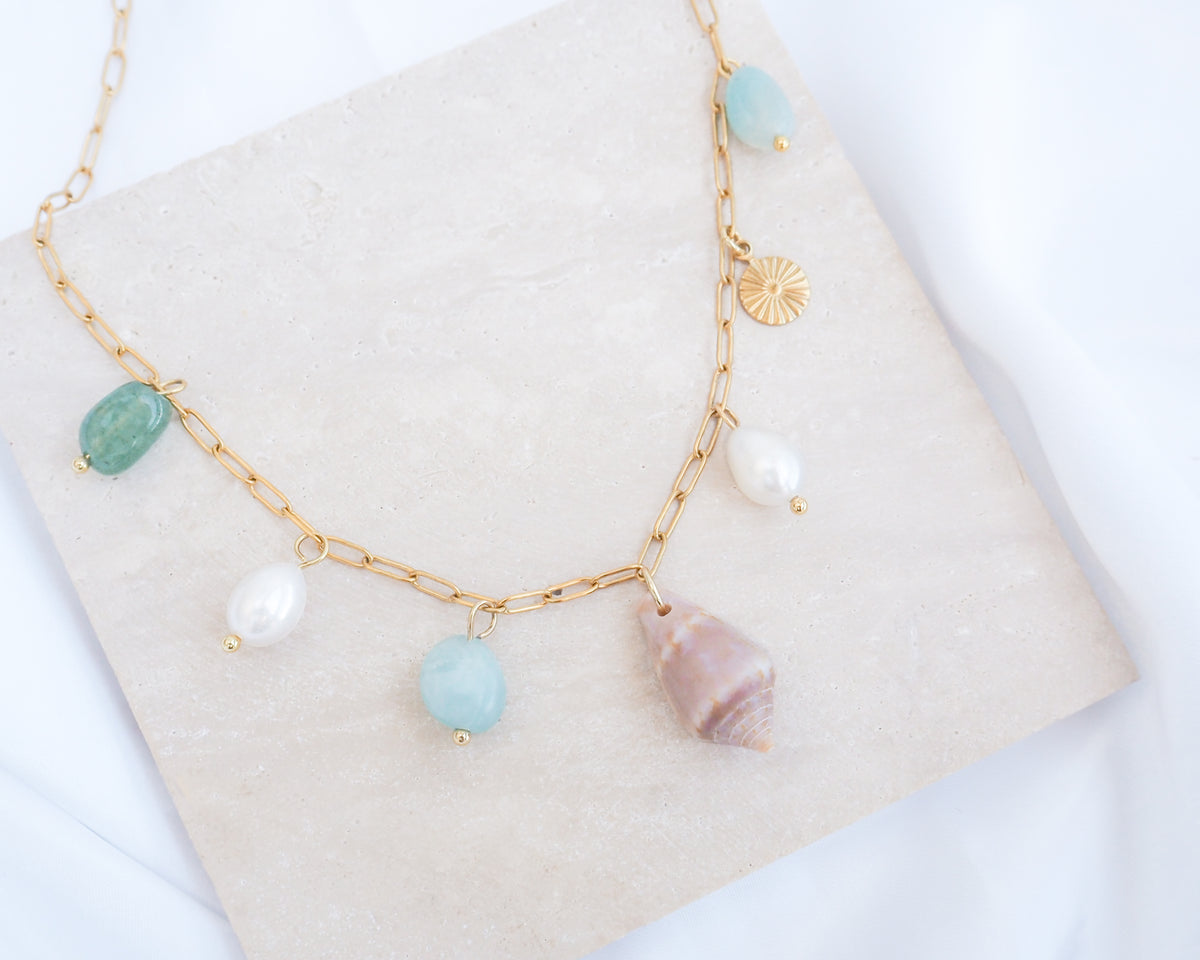 Close up of a gold charm necklace with a cone shell, ivory freshwater pearls and green aventurine gemstone beads displayed on a light grey stone. Made with stainless steel chain. The shell is a Mediterranean Cone Shell handpicked from the beaches of Portugal. 
