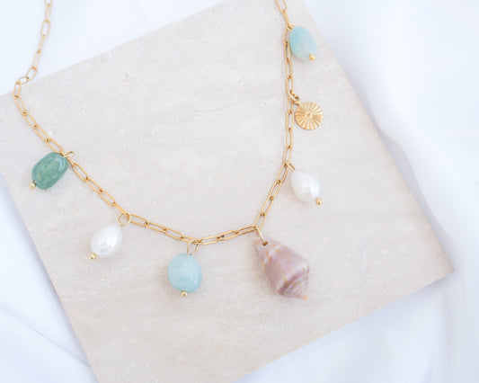 Close up of a gold charm necklace with a cone shell, ivory freshwater pearls and green aventurine gemstone beads displayed on a light grey stone. Made with stainless steel chain. The shell is a Mediterranean Cone Shell handpicked from the beaches of Portugal. 