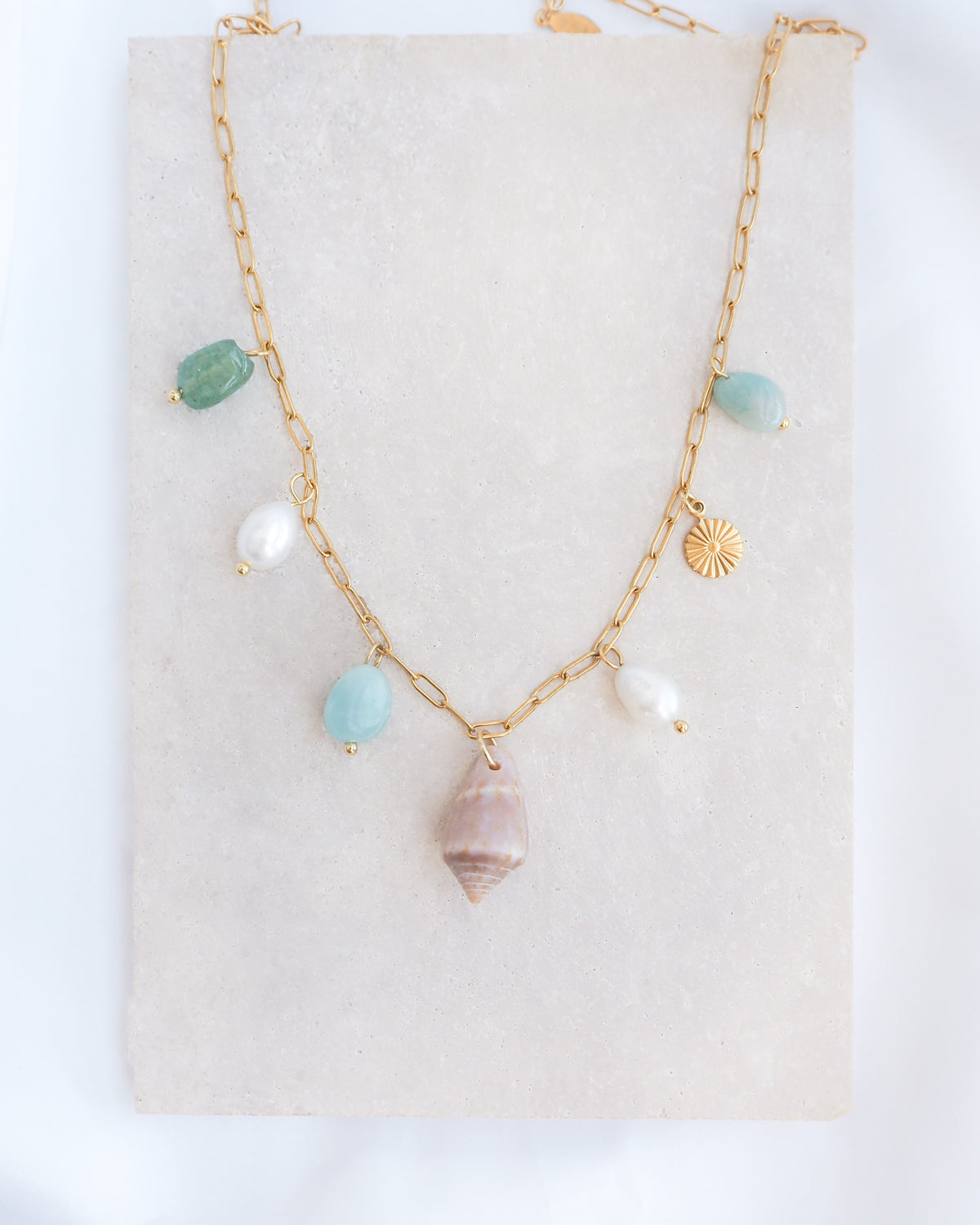 A gold charm necklace with a cone shell, ivory freshwater pearls and green aventurine gemstone beads displayed on a light grey stone. Made with stainless steel chain. The shell is a Mediterranean Cone Shell handpicked from the beaches of Portugal. 