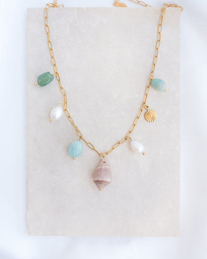 A gold charm necklace with a cone shell, ivory freshwater pearls and green aventurine gemstone beads displayed on a light grey stone. Made with stainless steel chain. The shell is a Mediterranean Cone Shell handpicked from the beaches of Portugal. 