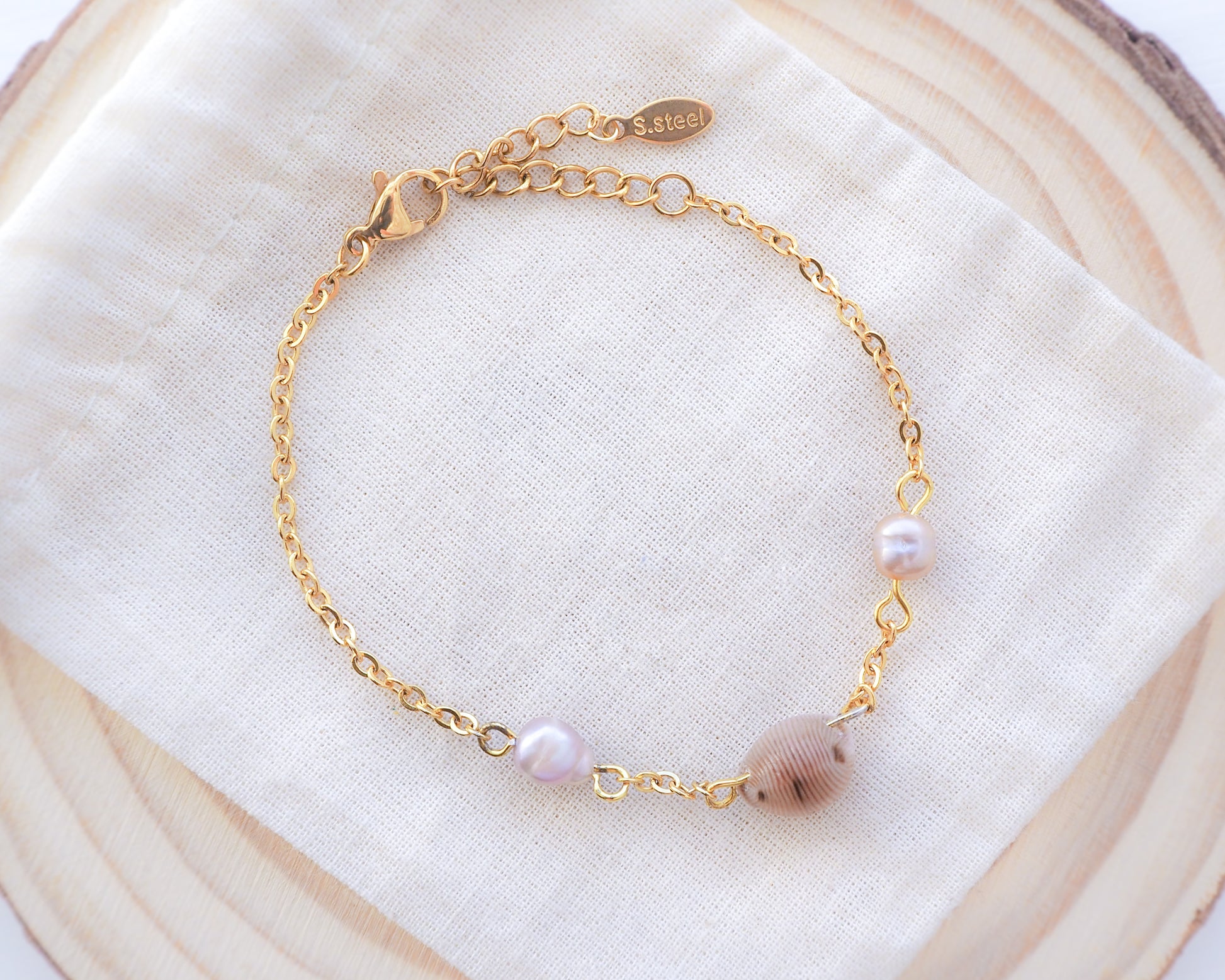 Backside of cowrie shell pearl bracelet on cotton displayed. Handpicked European cowrie shell from Portugal set on a 18k gold stainless steel bracelet chain and adorned with 2 freshwater peals that are pink-purple in color. The shell is about 8 mm and white in color. 