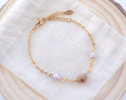 Backside of cowrie shell pearl bracelet on cotton displayed. Handpicked European cowrie shell from Portugal set on a 18k gold stainless steel bracelet chain and adorned with 2 freshwater peals that are pink-purple in color. The shell is about 8 mm and white in color. 