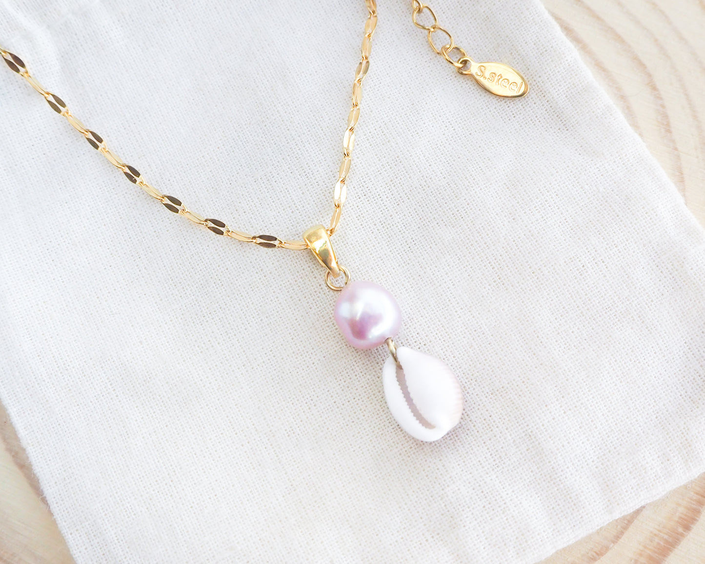 Delicate necklace with a pink pearl and shell pendant on a gold chain against a white fabric background.