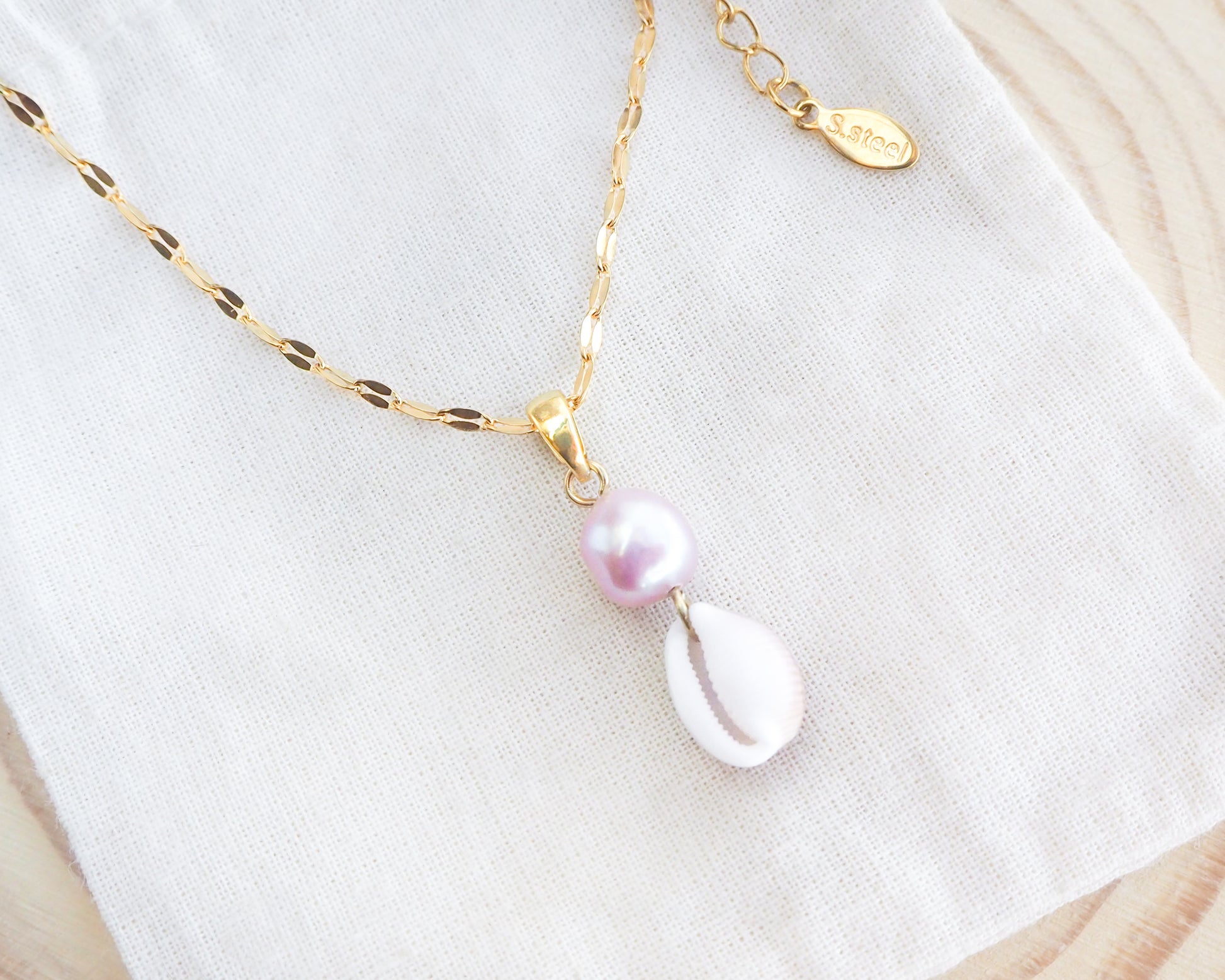 Delicate necklace with a pink pearl and shell pendant on a gold chain against a white fabric background.