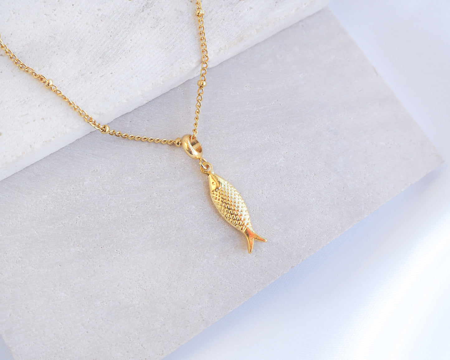 Gold fish-shaped pendant on a chain against a light background. Gold Pisces Fish pendant necklace on a marble surface. Made with 18k gold stainless steel. 