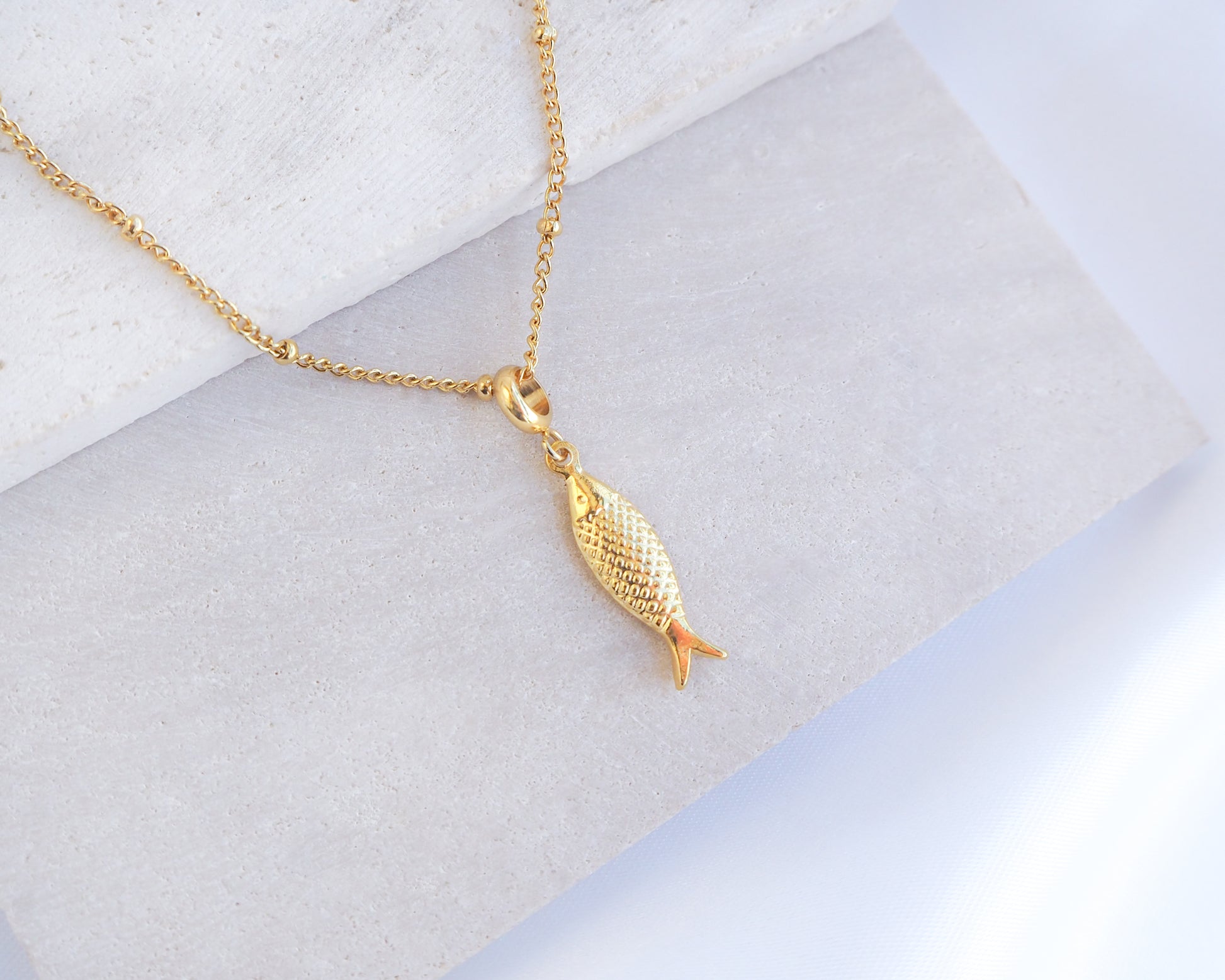 Gold fish-shaped pendant on a chain against a light background. Gold Pisces Fish pendant necklace on a marble surface. Made with 18k gold stainless steel. 