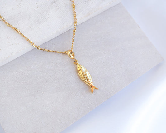 Gold fish-shaped pendant on a chain against a light background. Gold Pisces Fish pendant necklace on a marble surface. Made with 18k gold stainless steel. 
