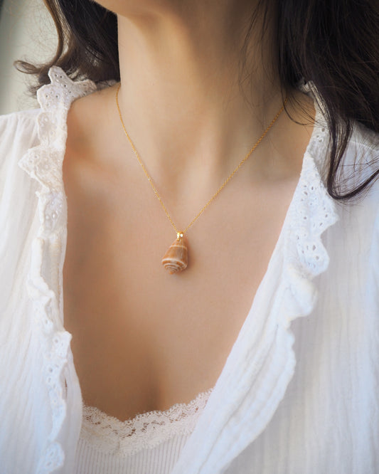 Woman wearing a gold necklace with a mediterranean cone shell pendant, dressed in a white lace top. The shell is orange brown in color and has a cone shape. The shell was collected from the beach of Portugal. 