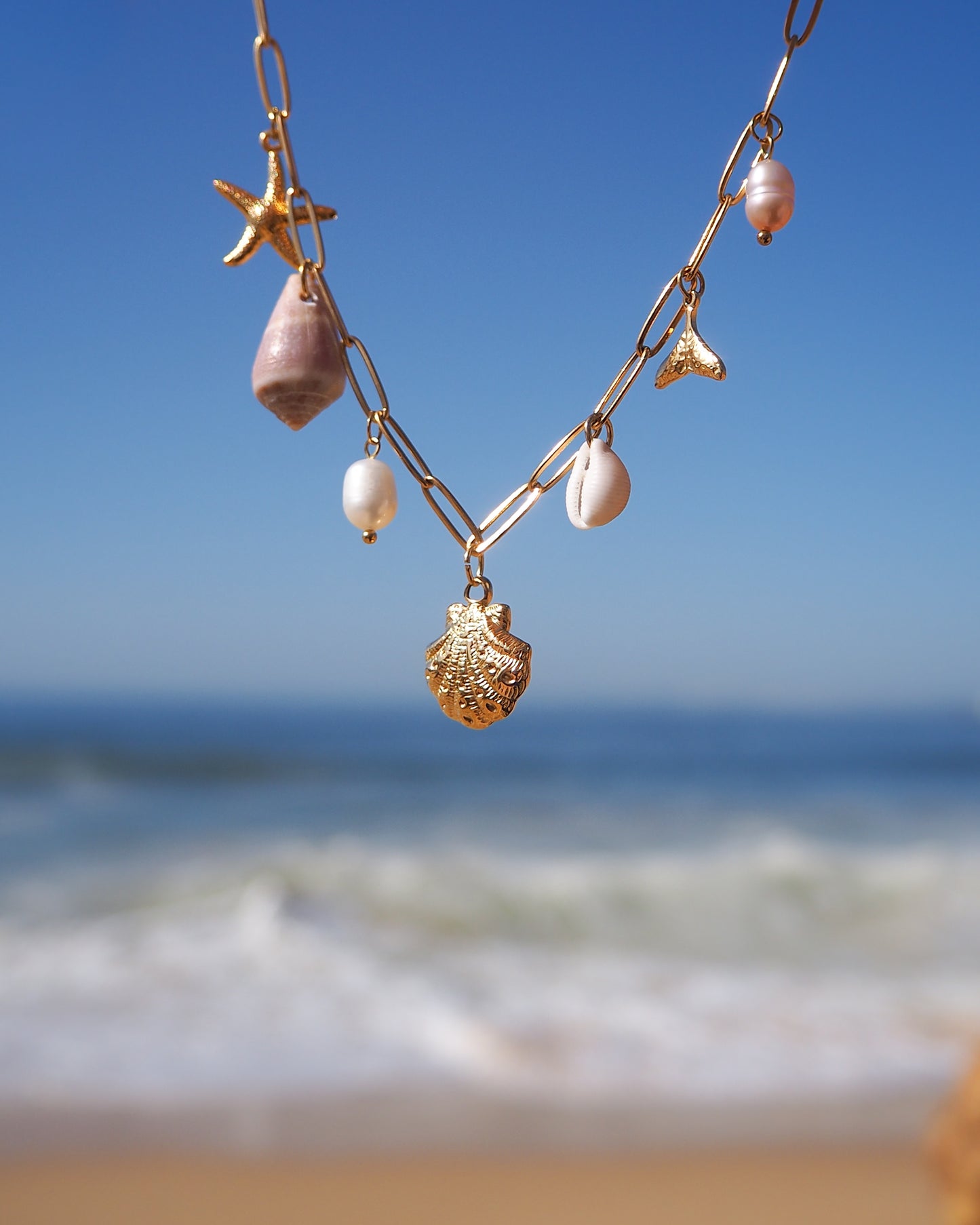 Gold Ocean Charm Necklace on neck with freshwater pearls, cone shell, cowrie shell, starfish sea star and whale tail pendants with ocean background. 