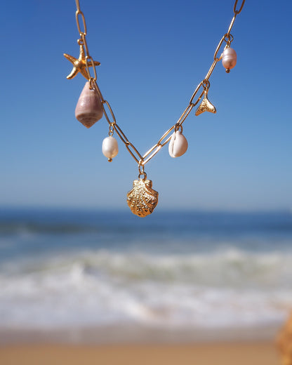 Gold Ocean Charm Necklace on neck with freshwater pearls, cone shell, cowrie shell, starfish sea star and whale tail pendants with ocean background. 