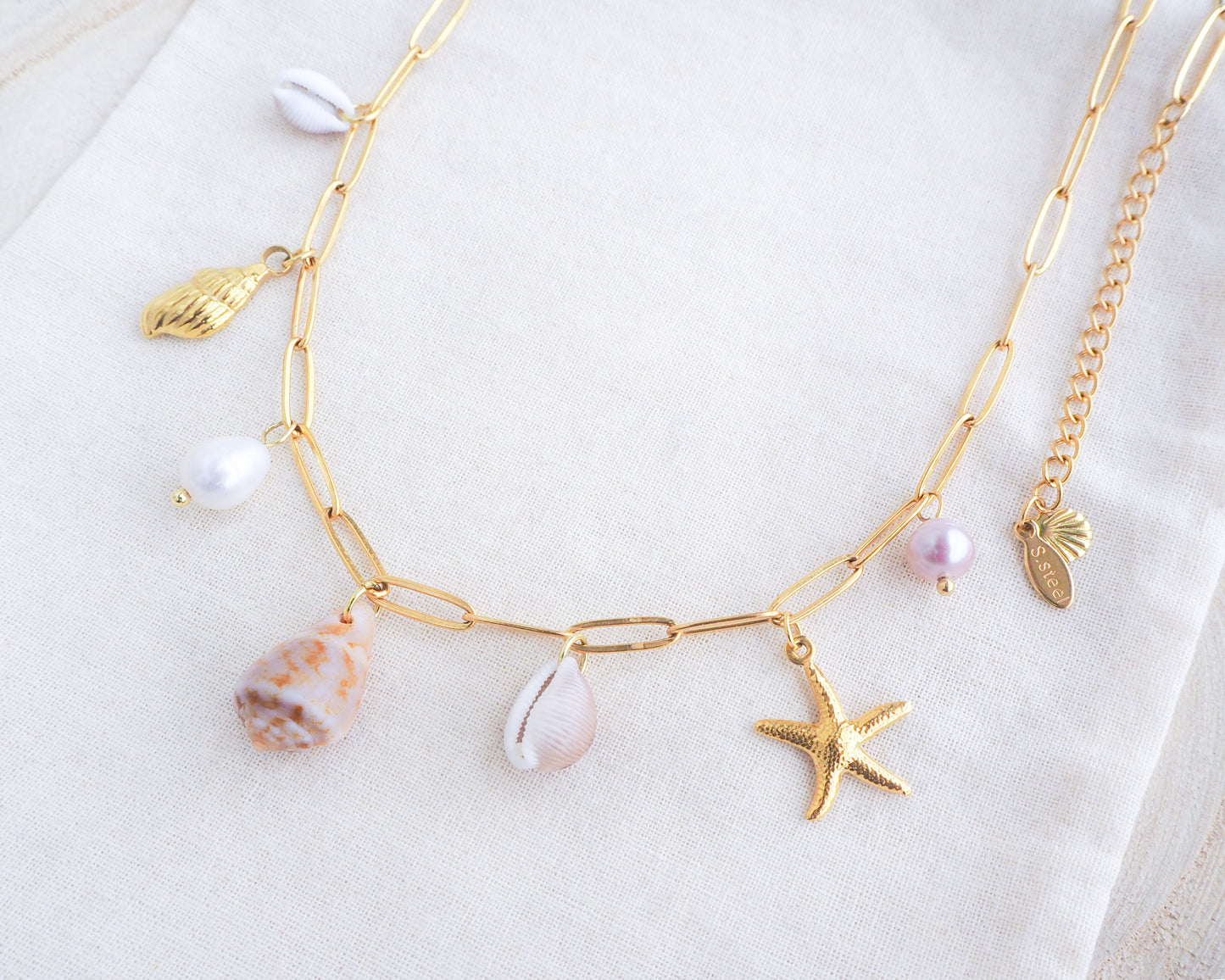Side view of Gold Ocean Shell Charm Necklace on display tiny cone and cowrie shells, freshwater pearls, starfish sea star and whale tail pendants. Made with a 18k gold stainless steel chain. The shells are handpicked from Portugal. 