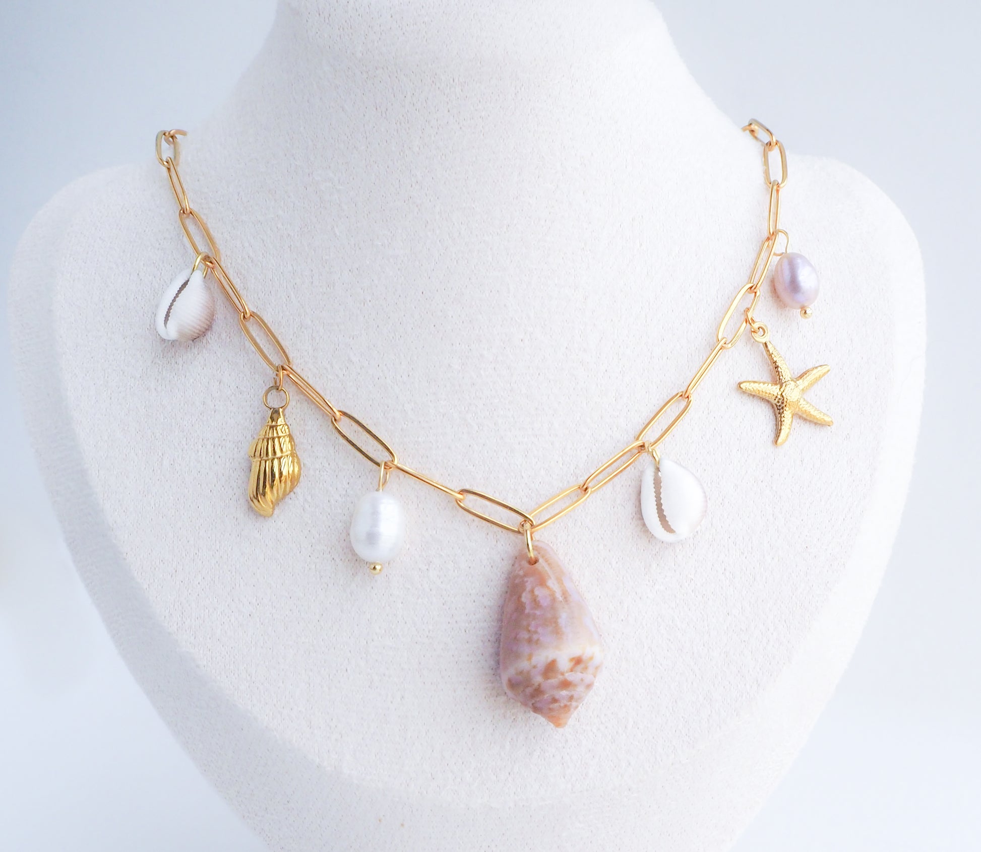 Gold necklace with various charms on a mannequin bust against a white background. Gold Ocean Shell Charm Necklace on mannequin, with cone and cowrie shells, freshwater pearls, starfish sea star shell pendants. 