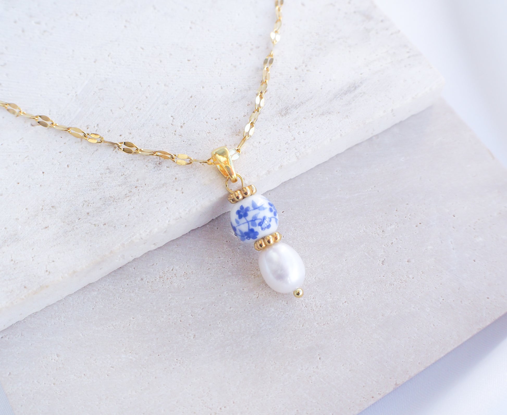 Azulejo Ceramic Tile Pearl Gold Necklace from Portugal. Close up of Gold Azulejo Pearl Necklace from Portugal, Lisbon Tile Jewelry. Made with premium 18k gold stainless steel chain, a ceramic bead and white freshwater pearl. The ceramic bead is showing a flower pattern with white and blue.