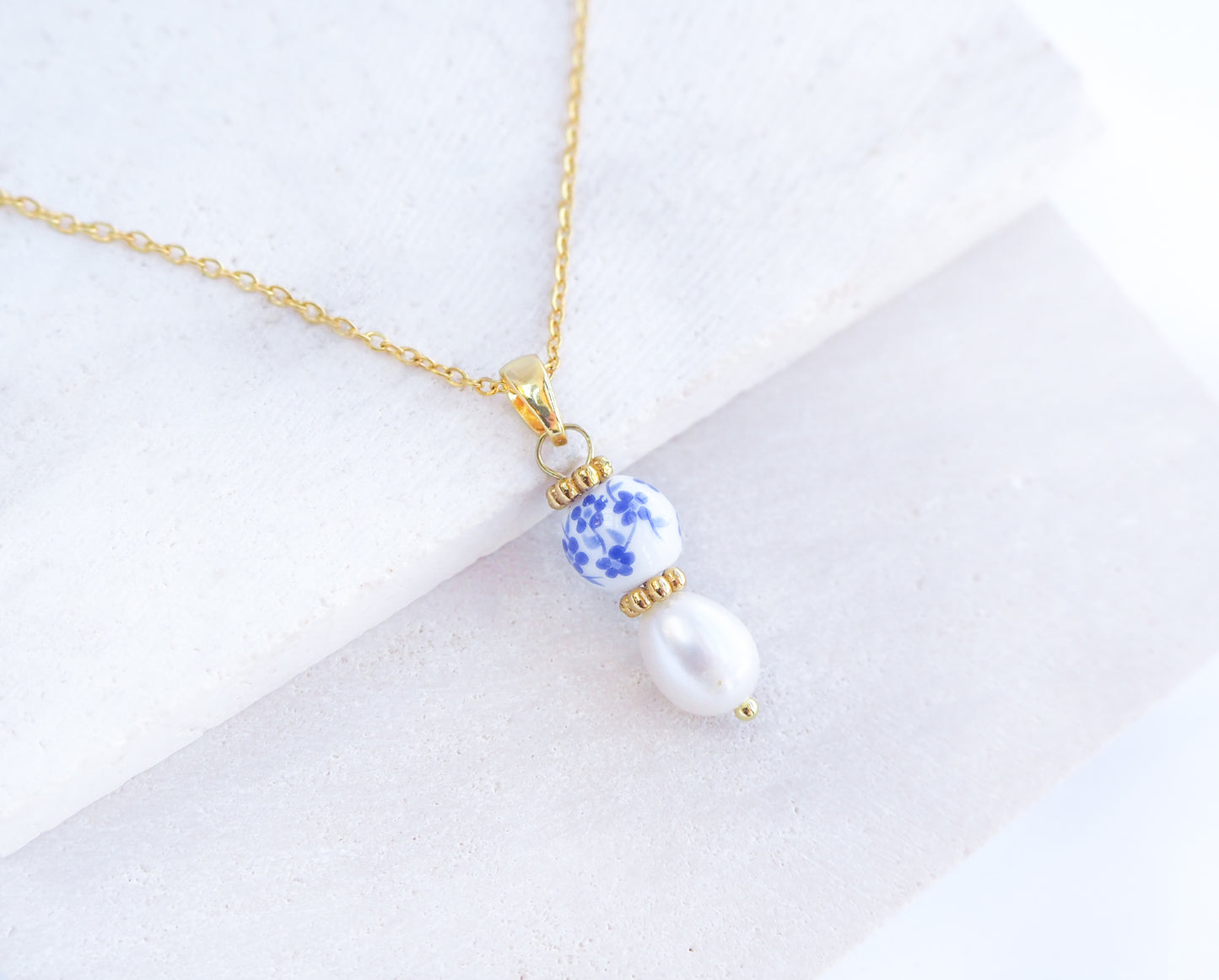 Close up of Gold Azulejo Pearl Necklace from Portugal, Lisbon Tile Jewelry. Made with premium 18k gold stainless steel chain, a ceramic bead and white freshwater pearl. The ceramic bead is showing a flower pattern with white and blue.