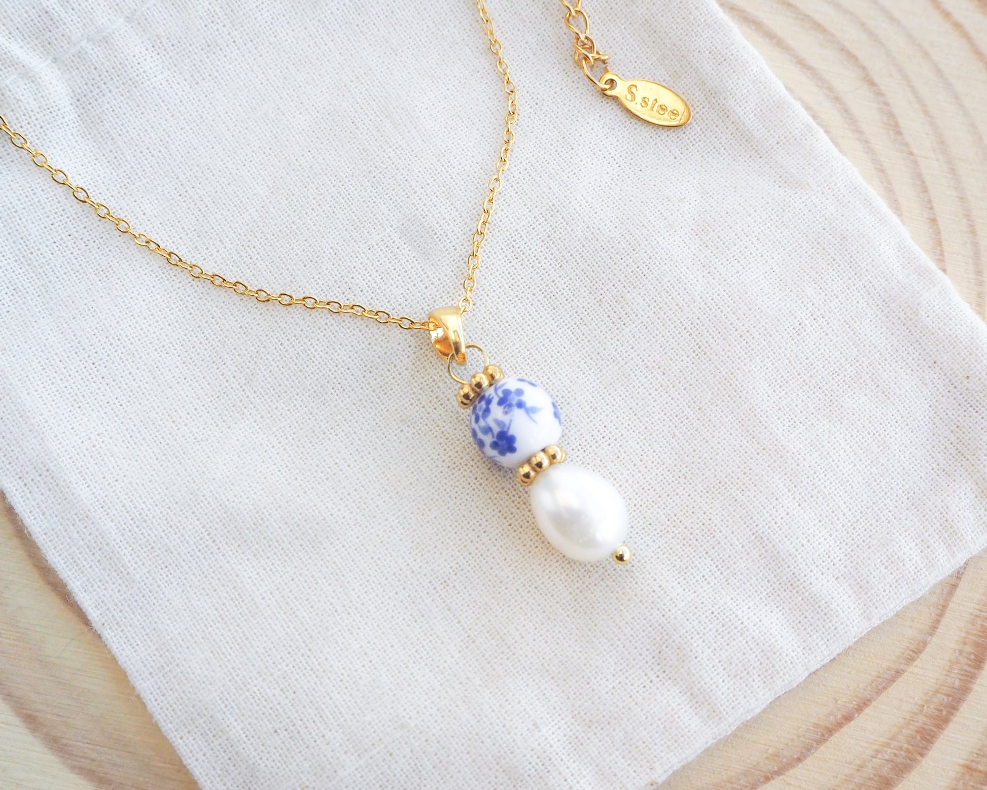 Close up of Gold Azulejo Pearl Necklace from Portugal on display, Lisbon Tile Jewelry. Made with premium 18k gold stainless steel chain, a ceramic bead and white freshwater pearl. The ceramic bead is showing a flower pattern with white and blue.