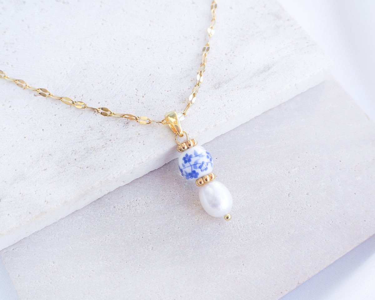 Azulejo Ceramic Tile Pearl Gold Necklace from Portugal. Close up of Gold Azulejo Pearl Necklace from Portugal, Lisbon Tile Jewelry. Made with premium 18k gold stainless steel chain, a ceramic bead and white freshwater pearl. The ceramic bead is showing a flower pattern with white and blue.