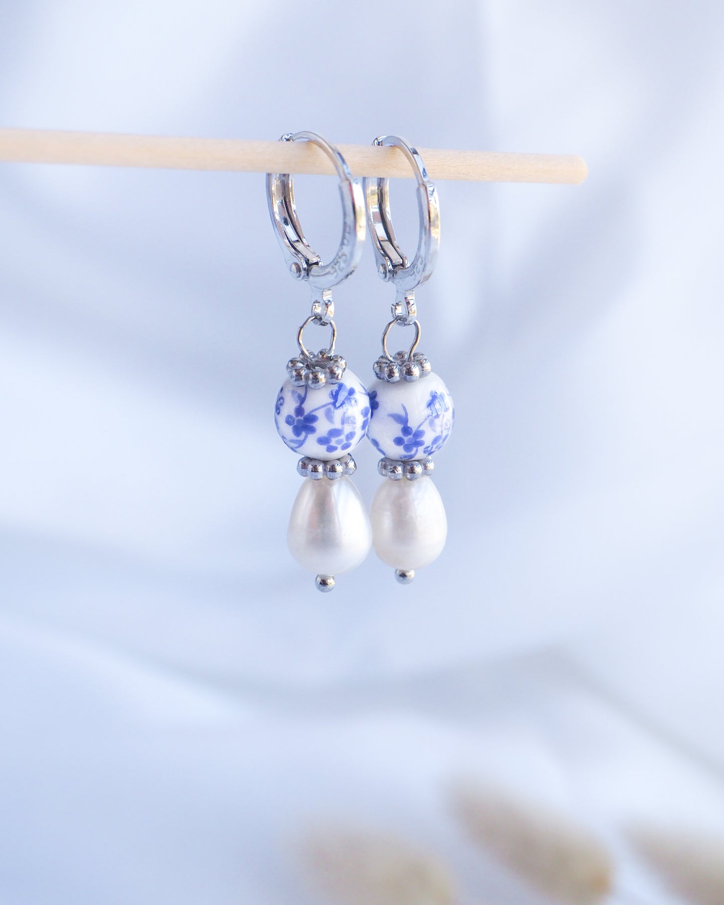 A pair of silver hoop earrings with a white pearl and a blue and white ceramic bead dangle. The ceramic bead has a floral pattern.