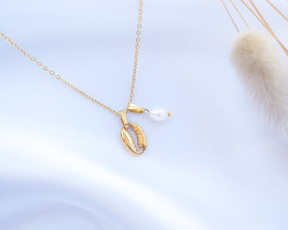 Gold necklace with a shell pendant and pearl on a light blue background. Gold Cowrie Shell Necklace with Pearl on display