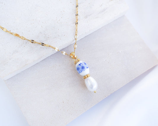 A gold chain necklace with a pendant featuring a blue and white patterned bead and a white pearl drop, displayed on a light gray stone block. Azulejo Flower Ceramic Tile Pearl Gold Necklace from Portugal 