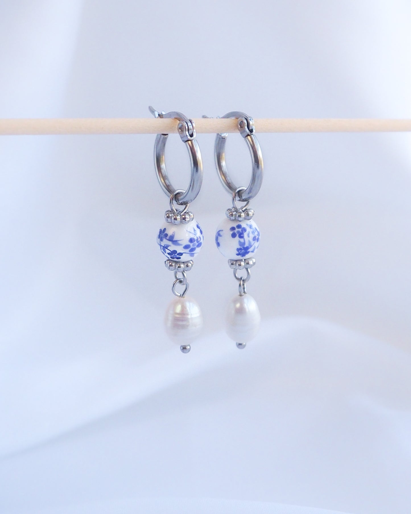 Pair of hoop earrings with floral beads and pearls on a white background. A pair of silver hoop earrings with a blue and white patterned bead and a white pearl dangling from each hoop.