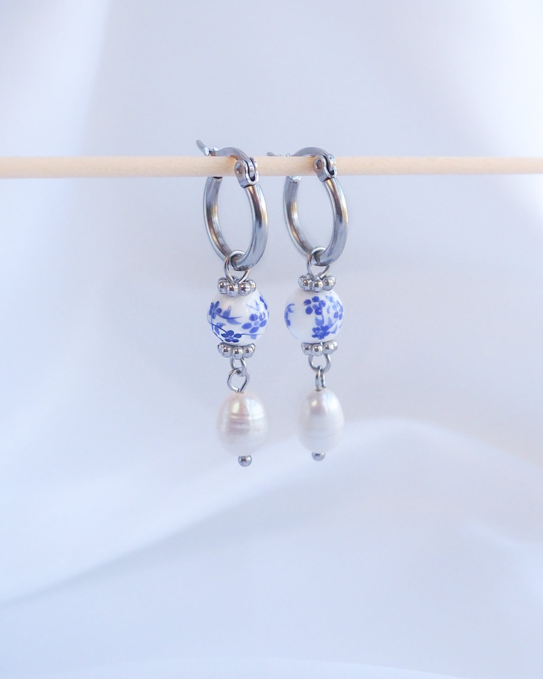 Pair of hoop earrings with floral beads and pearls on a white background. A pair of silver hoop earrings with a blue and white patterned bead and a white pearl dangling from each hoop.