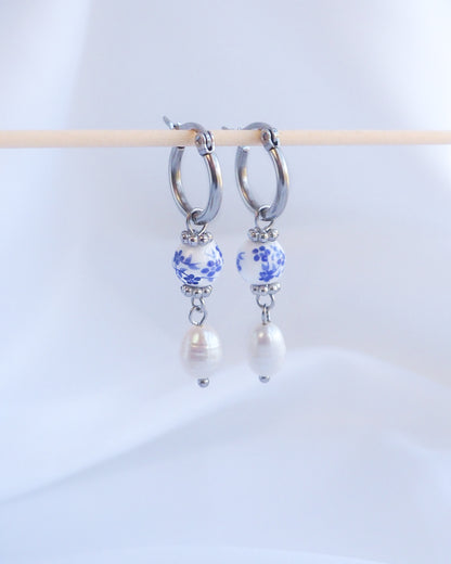 Pair of hoop earrings with floral beads and pearls on a white background. A pair of silver hoop earrings with a blue and white patterned bead and a white pearl dangling from each hoop.