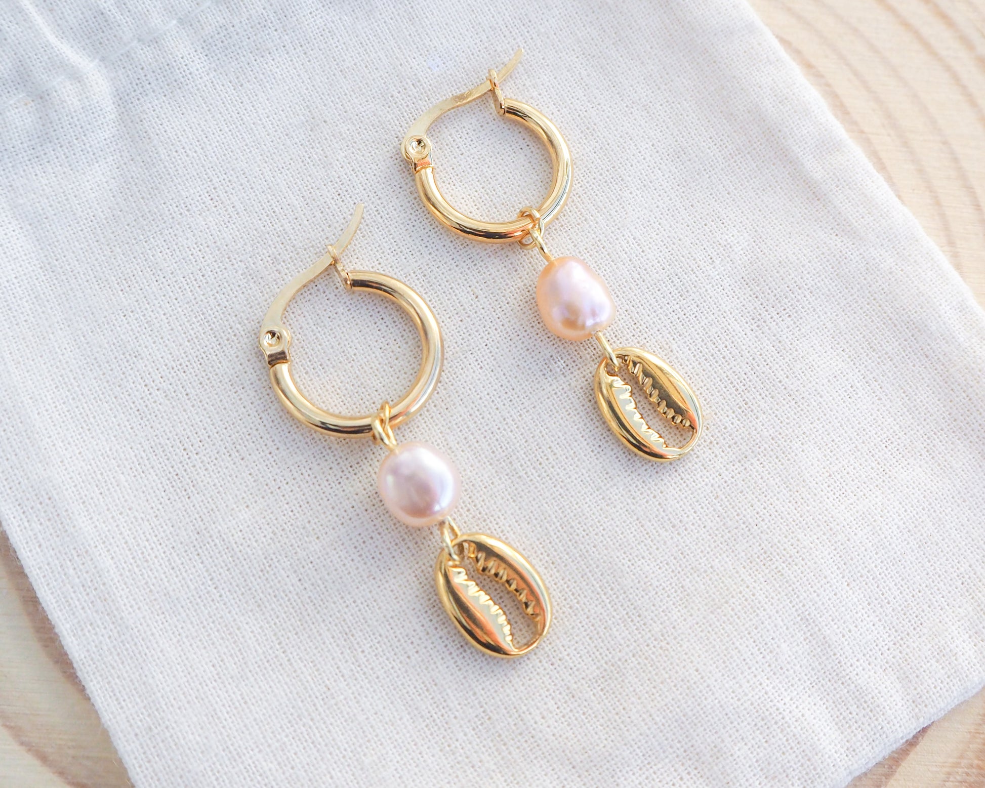 Gold hoop earrings with pearls on a white cloth background. Gold Hoop Earrings with Cowrie Shells and Pearls on cotton displayed. Made with premium 14k and 18k gold stainless steel and pink freshwater pearl beads. 