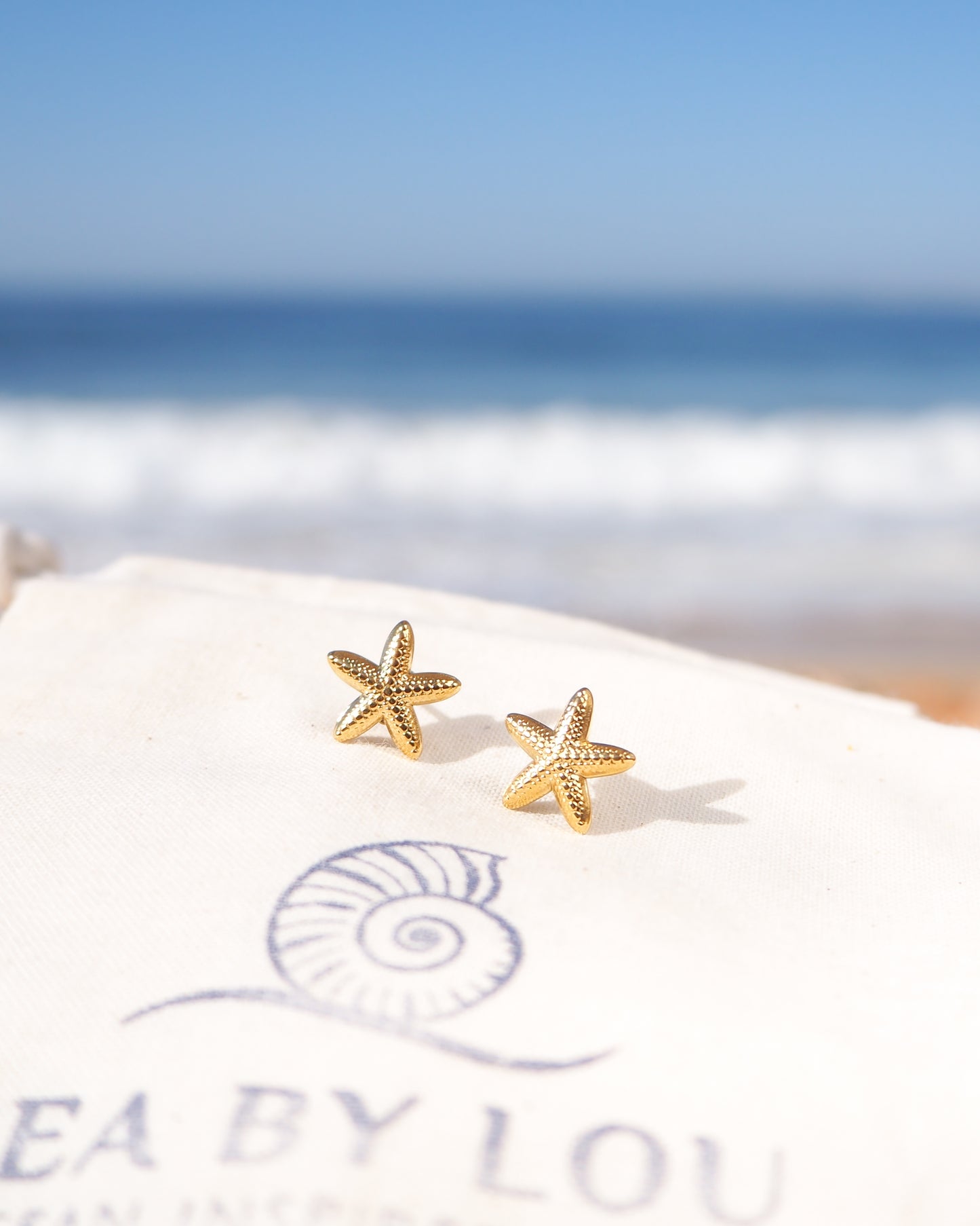 Gold Starfish Stud Earrings displayed on cotton with ocean background.