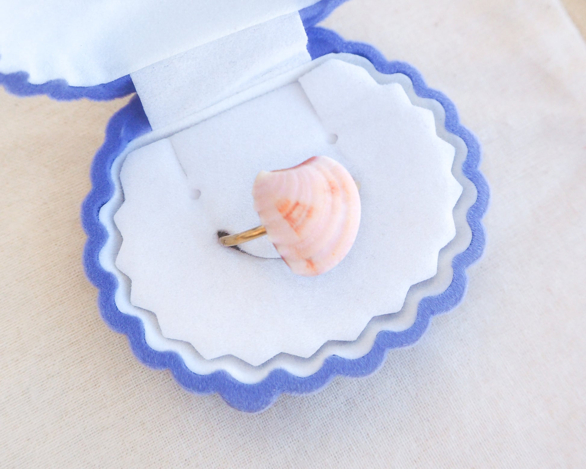 A seashell ring with a gold band sits in a blue and white scalloped jewellery box. The ring features a light pink shell with orange, pink and white markings. The jewellery box is open, displaying the ring against a white interior.