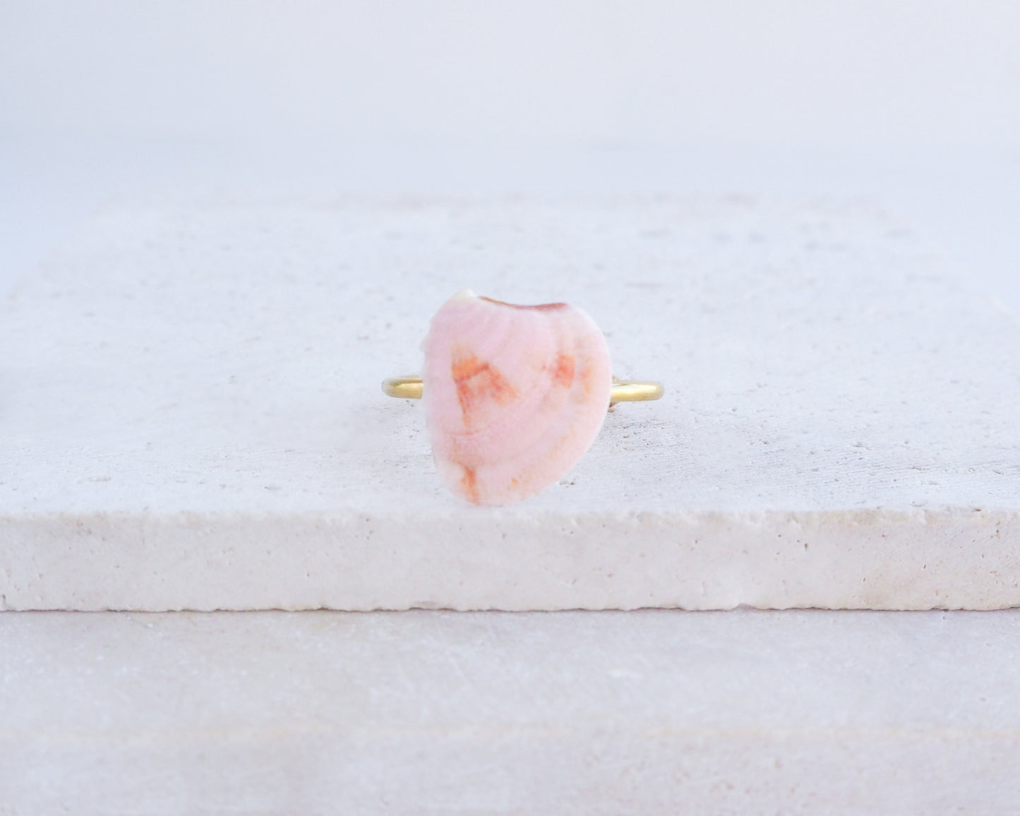 A soft pink seashell ring made with a handpicked venus shell from Portugal, displaying orange and white patterns, set on a thin, gold-coloured band. The gemstone is approximately 2 cm wide.