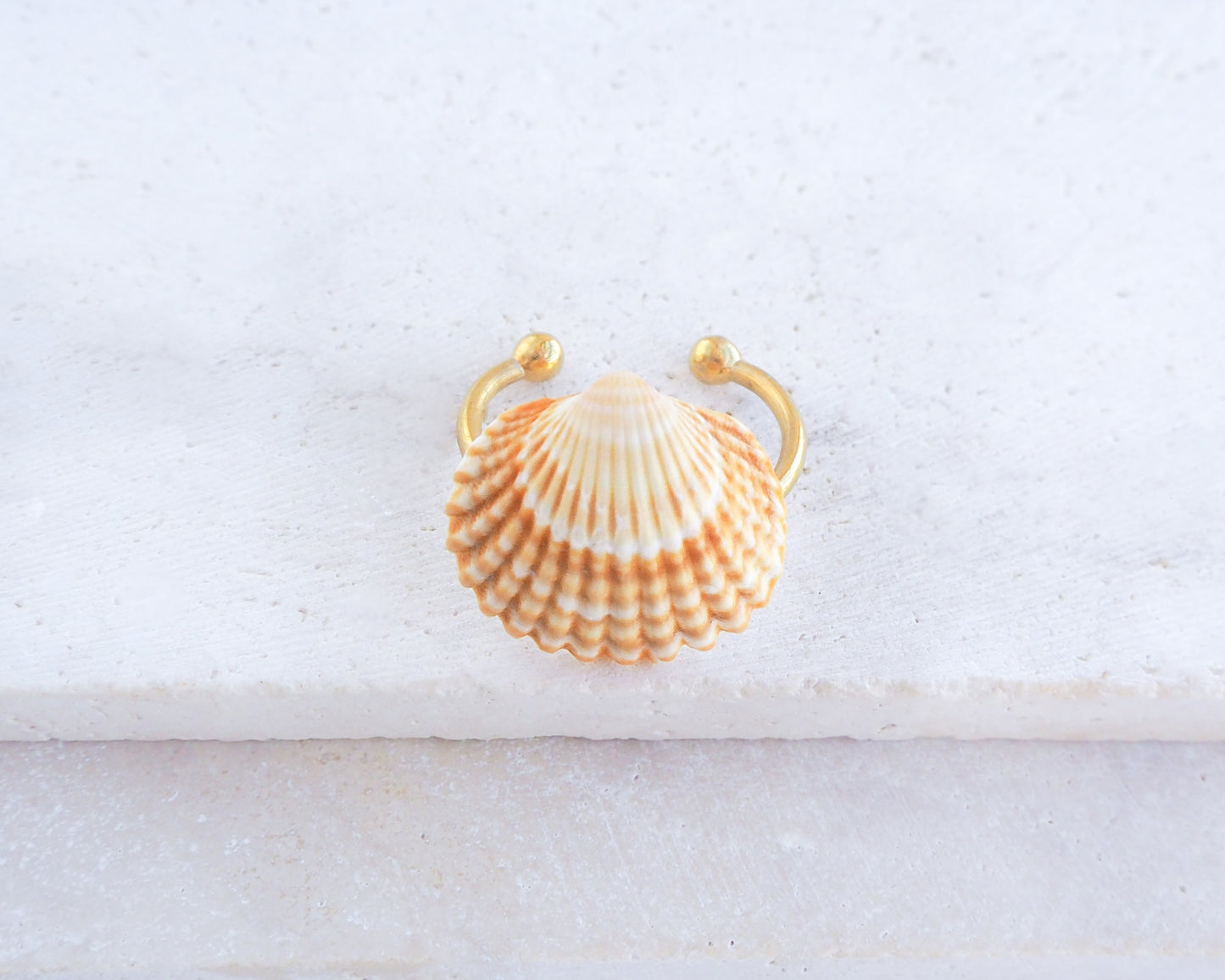 A seashell ring with handpicked mediterranean cockle shell from portugal and 18k gold stainless steel ring. The shell is cream and orange, with a ribbed texture. The hardware is a decorative accent for coastal or nautical-themed furniture.