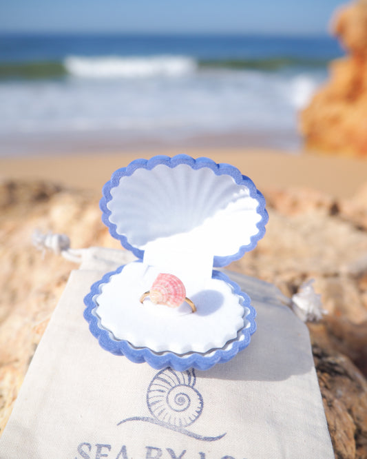 Ring in a shell-shaped box on a beach with 'SEA BY' branding. Pink White Venus Shell Ring from Portugal displayed on cotton with ocean background, handpicked real shell from Portugal