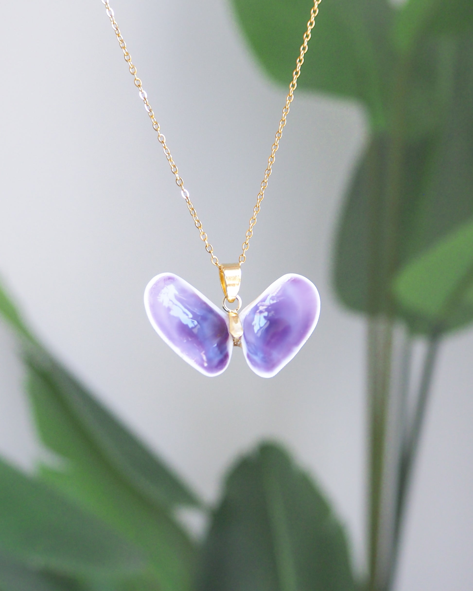 A gold chain necklace with a butterfly pendant. The pendant is made of two purple, shell-shaped pieces with a white border, forming butterfly wings. The necklace is set against a soft, blurred background.