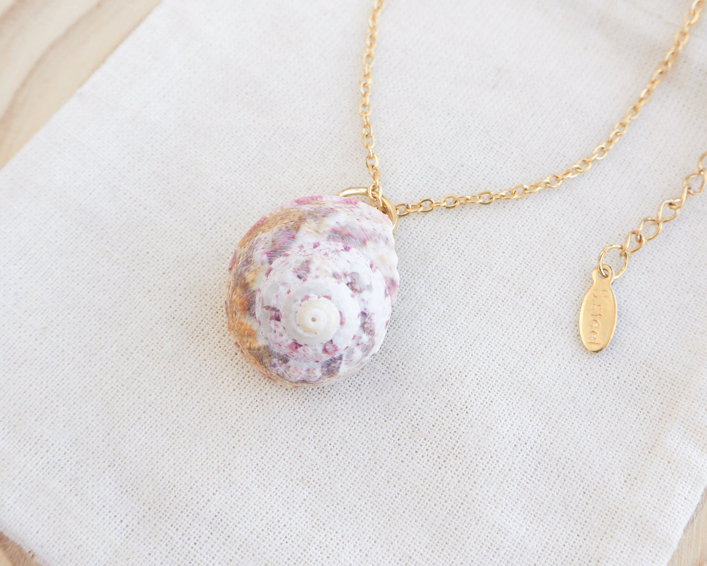 Sunny Pink White Orange Magus Top Shell Gold Necklace on display side view. A gold-coloured chain necklace with a seashell pendant. The pendant is a mix of pink, white, and gold. The necklace is displayed on a white cloth.